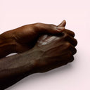 Two dark-skinned hands applying Aesop Eleos Aromatique Hand Balm cream on a light pink background