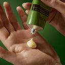 Green tube of Aesop Geranium Leaf Body Balm 100ml being squeezed onto a hand against a green background