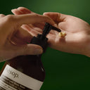Person applying Aesop Geranium Leaf Body Balm 500ml to their hand against a green background