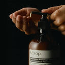 Person holding a brown Aesop Resurrection Aromatique Hand Balm 500ml bottle with a pump against a black background