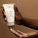 Hand holding Aveda hand relief cream against a brown background