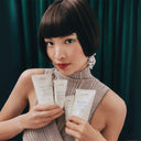 Woman holding Aveda skincare products against a green curtain background