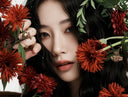 A close-up portrait of a woman with dark hair surrounded by red flowers and green leaves, with one hand holding a flower stem near her face.