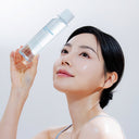 Woman holding a clear blue bottle of Beauty of Majesty BOM Hyaluronic Acid Moisture Toner 200ml with a white cap against a light background