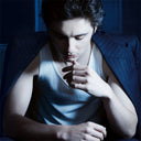 Timothée Chalamet in a white tank top and navy pinstriped suit jacket sitting in a dimly lit room with a blue hue.