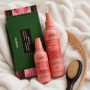 BEAUTE KNOWS Aveda Nutriplenish leave-in conditioner and overnight serum bottles arranged on a white plush towel beside a green product box and a wooden hairbrush.