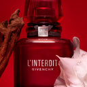 BEAUTE KNOWS Red Givenchy L'interdit Rouge Eau De Parfum 80ml perfume bottle with a white flower and wooden object on a red background