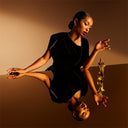 Woman in a black dress sitting on a reflective surface with a bottle of Jean Paul Gaultier Gaultier Divine Elixir Parfum 100ml