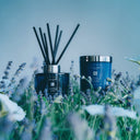 blue Jo Malone London Lavender & Moonflower diffuser with black reeds on the left and Jo Malone London Lavender & Moonflower scented candle on the right seating in a field of lavender flowers with blurred background