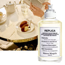 Maison Margiela Replica Afternoon Delight Eau de Toilette 100ml perfume bottle with a blurred background of a person reading a book and a tea set.