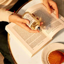 Hand holding a bottle of Maison Margiela Replica Afternoon Delight Eau de Toilette 100ml perfume over an open book with a plate of food on a table.