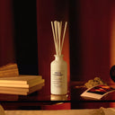 WhiteM aison Margiela Replica By the Fireplace Scented diffuser bottle labeled 'REPLICA' on a wooden surface with books and a magazine in the background.