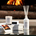 Maison Margiela Replica By the Fireplace Candle, diffuser, and photo book on a table with a fireplace in the background
