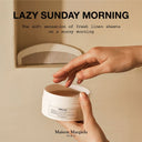 BEAUTE KNOWS Hand holding a Maison Margiela Replica Lazy Sunday Morning Body Scrub container with 'Maison Margiela' branding, text 'Lazy Sunday Morning' on a beige background.