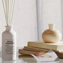 White Maison Margiela Replica Lazy Sunday Scented diffuser bottle labeled 'REPLICA' with a wooden vase and books on a light background