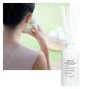 White Maison Margiela Replica Matcha Meditation Scented Diffuser bottle with 'REPLICA' labelling next to a blurred person drinking matcha tea.