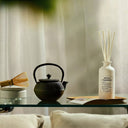 Tea-making set with black teapot, wooden whisk, and Maison Margiela Replica Matcha Meditation Scented Diffuser on a glass table.