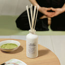 White Maison Margiela Replica Matcha Meditation Scented Diffuser 185ml bottle with reeds on a wooden table, blurred person in the background