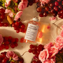 Maison Margiela Replica On A Date Eau de Toilette perfume bottle surrounded by pink flowers and red grapes on a light background