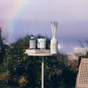 Table with Maison Margiela Replica When the Rain Stops Perfume,Scented Diffuser and candle against a window with a rainbow outside