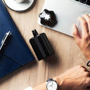 Black Montblanc Emblem Eau de Toilette 100ml perfume bottle on a wooden desk with a hand wearing a watch, a laptop, and a notebook.