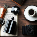 Collection of items including Montblanc Emblem Eau de Toilette 100ml perfume bottle, a watch, camera, coffee, and book on a wooden surface