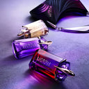 Three colorful Mugler Alien perfume bottles with a 'Mugler' bag in the background on a textured surface.