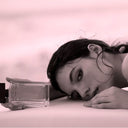 Woman lying on a pink surface with a clear Narciso Rodriguez For Her Musc Noir Eau de Parfum 100ml perfume bottle in front of her.