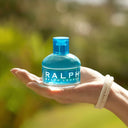Hand holding a Ralph Lauren Ralph Eau de Toilette 50ml blue bottle against a blurred green background