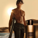 Man holding a surfboard with a Tom Ford Oud Minérale perfume bottle in a desert setting
