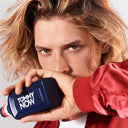 a blonde male model holding a Tommy Hilfiger Tommy Now Eau de Toilette 100ml bottle with a red jacket against a neutral background