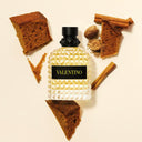 Valentino Uomo Born In Roma Yellow Dream Eau de Toilette 50ml perfume bottle surrounded by brown bread slices, nuts, and cinnamon sticks on a beige background