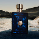 Blue Versace Dylan Blue Pour Homme Eau de Toilette 50ml perfume bottle with gold cap on a beach with mountains in the background