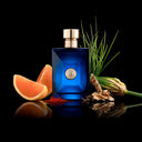 Blue Versace Dylan Blue Pour Homme Eau de Toilette 50ml perfume bottle with a silver cap on a reflective surface with orange slices, grass, and flowers.