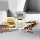 YSL Yves Saint Laurent L'Homme Eau de Toilette 100ml bottle with silver cap and hexagonal design, surrounded by a lemon and dried leaves on a light gray surface.